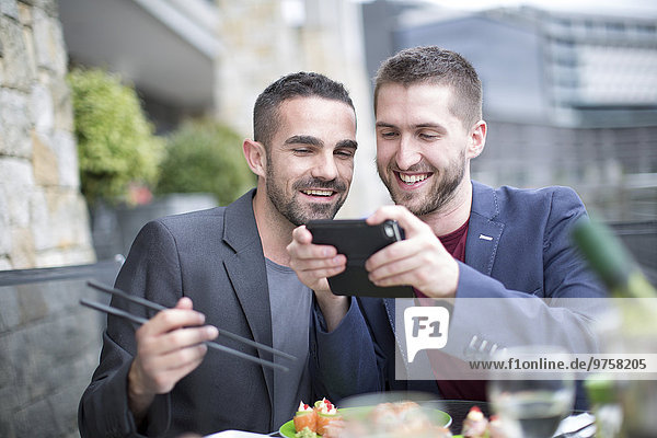 Schwules Paar isst Sushi und schaut sich das digitale Tablett im Außenrestaurant an.