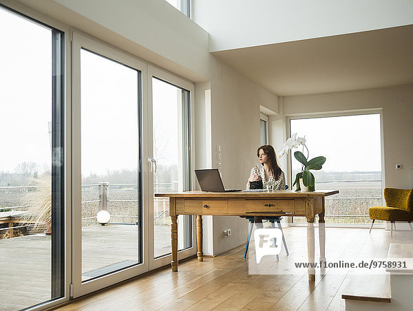 Young woman with laptop at wooden table looking out of window