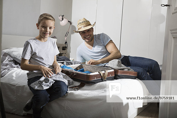 Father and son sitting on bed packing suitcase