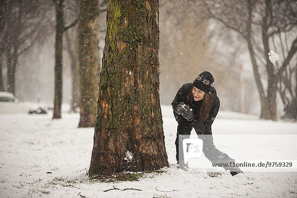 Junge Frau mit Schneeball versteckt sich hinter Baumstamm
