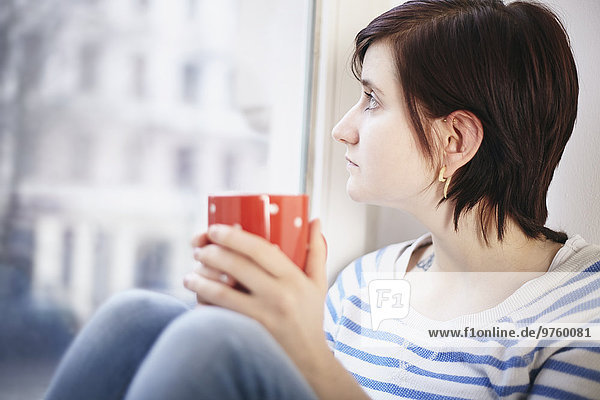 Junge Frau mit Tasse Kaffee durchs Fenster schauend