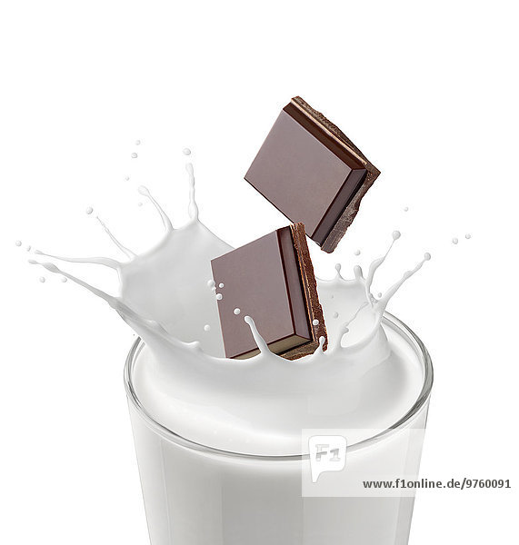 Zwei Stücke Schokolade fallen in ein Glas Milch vor weißem Hintergrund.