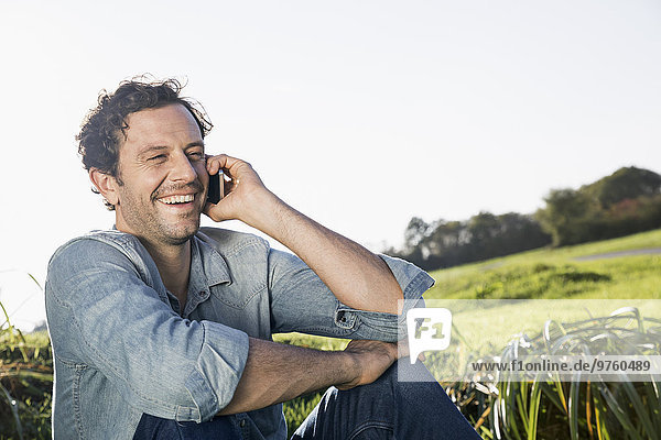 Laughing man sitting on a meadow telephoning with smartphone