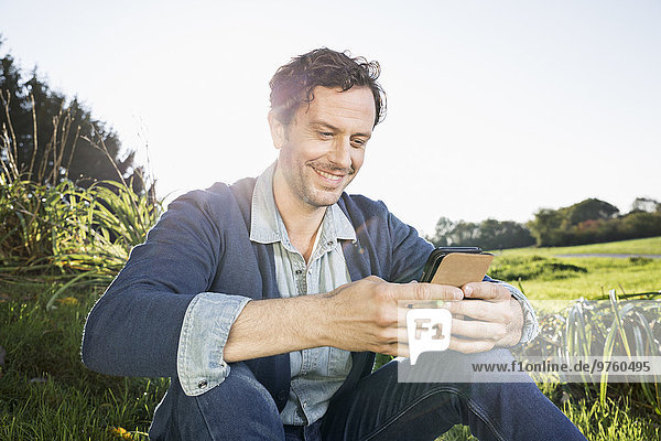 Smiling man sitting on a meadow reading SMS