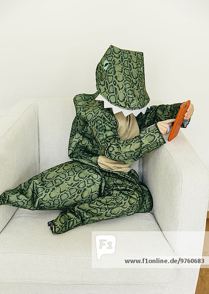 Kleiner Junge im Dinosaurierkostüm sitzend auf der Couch mit Smartphone