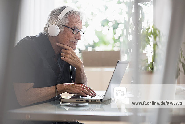Senior man sitting in home office wearing headphones and using laptop