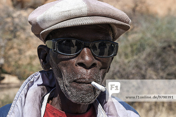 Einheimischer Mann mit Sonnenbrille und Mütze  raucht eine Zigarette  Portrait  bei Spitzkoppe  Namibia  Afrika