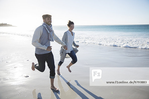 Junges Paar hält sich an den Händen und rennt am Strand.