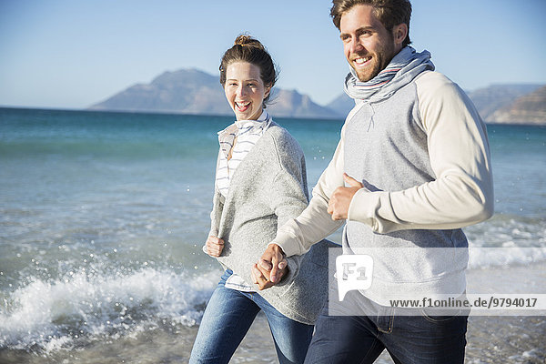 Junges Paar hält sich an den Händen und geht am Strand spazieren.
