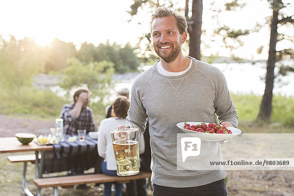 Glücklicher Mann mit Bierkrug und Erdbeeren mit Freunden am Picknicktisch im Hintergrund