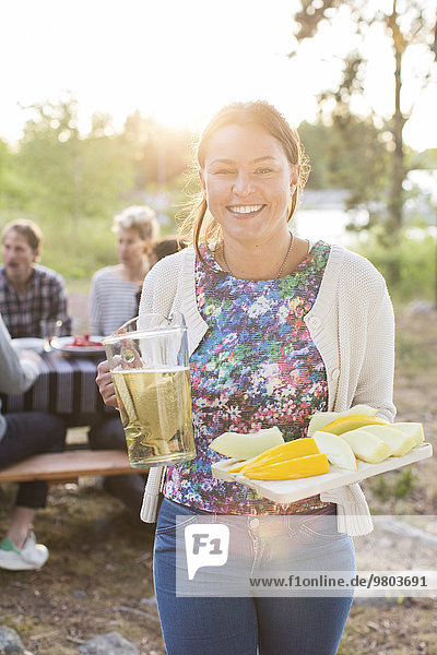 Porträt einer glücklichen Frau mit Bierkrug und Zuckermelonenscheiben mit Freunden am Picknicktisch im Hintergrund.