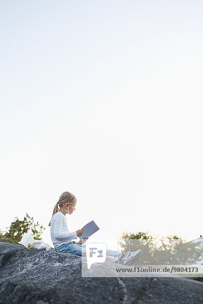 Volle Seitenansicht des Mädchens bei den Hausaufgaben  während es auf dem Felsen gegen den klaren Himmel sitzt.