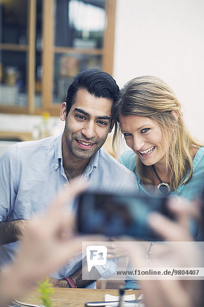 Freund fotografiert lächelndes Paar durch Smartphone im Cafe