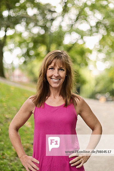 Portrait of happy fit mature woman standing with hands on hips at park
