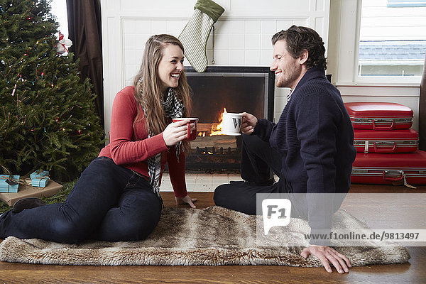 Young couple drinking coffee in front of log fire at christmas