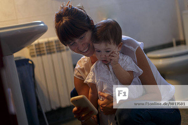 Mittlere erwachsene Frau und kleine Tochter mit Blick auf das Smartphone im Bad
