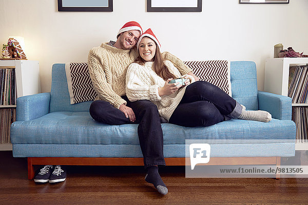 Porträt eines jungen Paares in Weihnachtsmützen auf Sofa mit Weihnachtsgeschenk