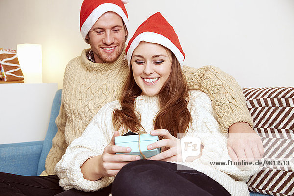 Junges Paar mit Weihnachtsmützen auf dem Sofa mit Blick auf das Weihnachtsgeschenk