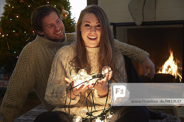 Young couple in living room holding illuminated christmas lights