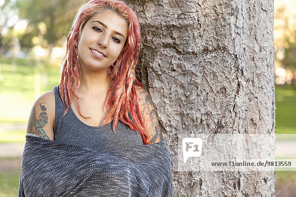 Young woman with pink dreadlocks leaning against tree trunk in park