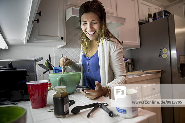 Frau bereitet Essen in der Küche zu  während sie das Rezept vom Smartphone liest.