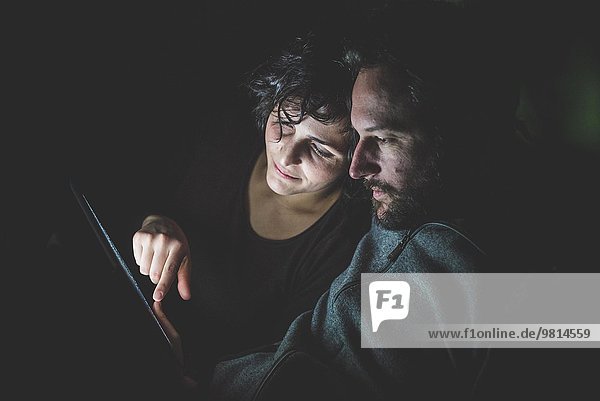 Paar sitzt in einem dunklen Raum und schaut auf ein digitales Tablett.