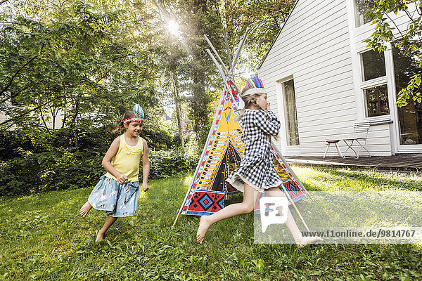 Two girls in native American headdress running around teepee in garden