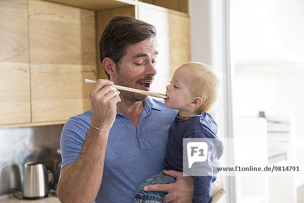 Man feeding toddler son with wooden spoon in kitchen