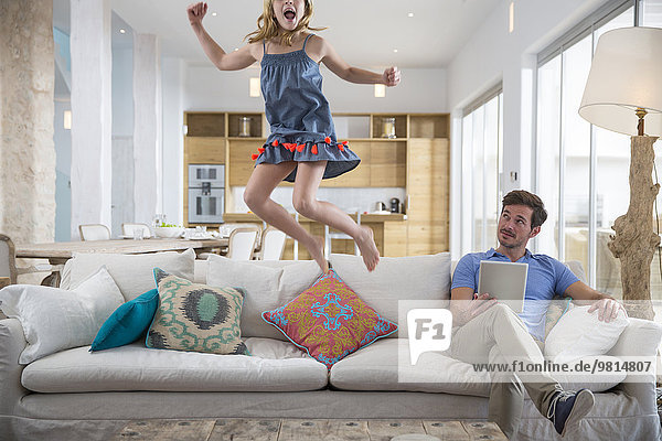 Mädchen springt in der Luft vom Wohnzimmersofa  während Vater ein digitales Tablett benutzt.