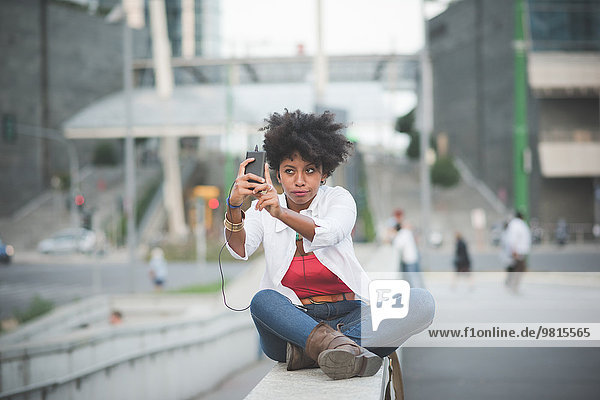 Junge Frau nimmt Smartphone Selfie  während sie auf der Wand des Einkaufszentrums sitzt.