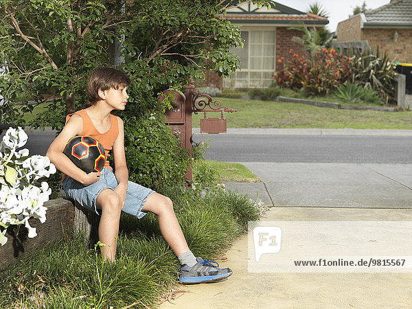 Mürrischer Junge sitzt auf einer Vorstadtmauer und hält einen Fußball.