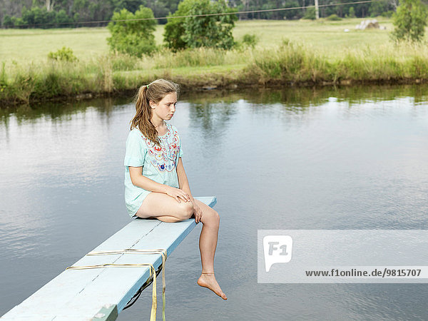 Teenage girl sitting on diving board over lake