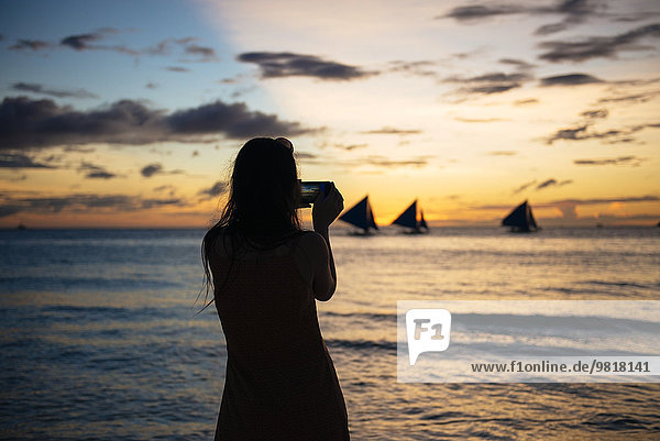 Philippinen  Boracay  Frau beim Fotografieren des Sonnenuntergangs mit Segelbooten