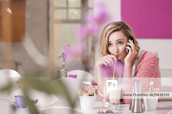 Lächelnde blonde Frau sitzt in einem Kaffee mit einem Glas Latte Macchiato beim Telefonieren.