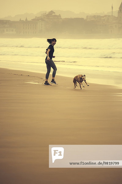 Spanien  Gijon  Frau beim Joggen am Strand  Blick auf den laufenden Hund