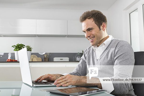 Smiling man sitting in kitchen using laptop