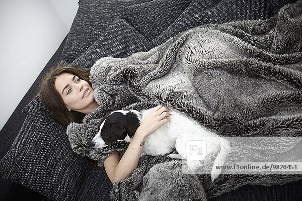 Young woman wrapped in fur blanket relaxing on couch with dog