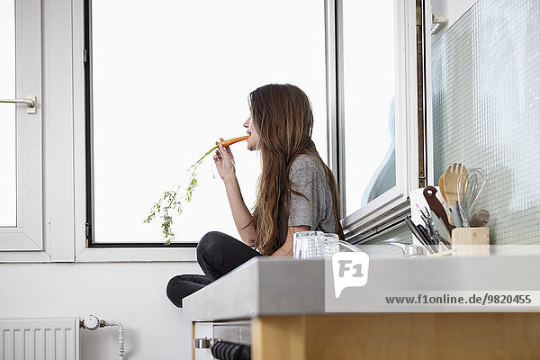 Junge Frau in der Küche sitzt am Fenster und isst Karotten.
