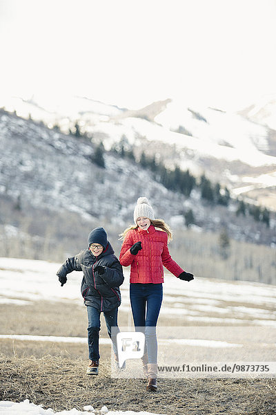 A brother and sister running across the grass together n winter.