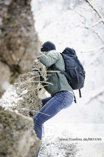 A man wearing a fleece jacket and hat,  carrying a rucksack,  climbing up a rocky cliff face.