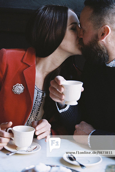 A couple seated side by side at a cafe table  kissing.