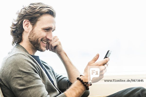 A bearded man sitting smiling and checking his phone.