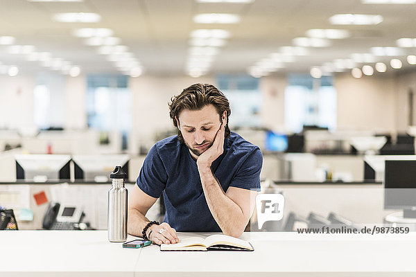 A man leaning on his elbow and looking at an open book on an office desk.