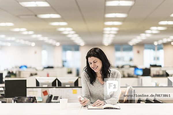 Eine Frau sitzt an einem Schreibtisch mit der Hand auf einem aufgeschlagenen Buch und schaut auf ihr Smartphone.