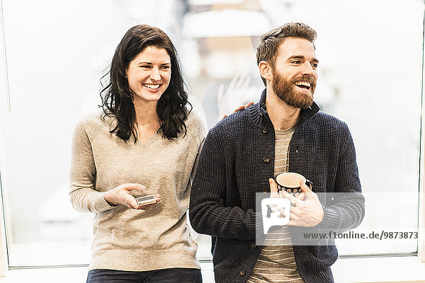 Eine Geschäftsfrau sitzt am Fenster  hält ein Smartphone in der Hand und unterhält sich mit einem Mann  der eine Kaffeetasse hält.