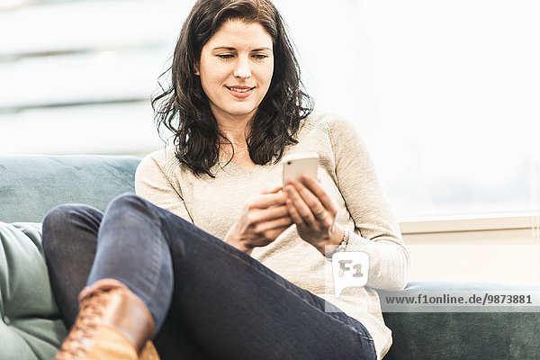 A woman seated with her feet up on a sofa  looking at her smart phone.