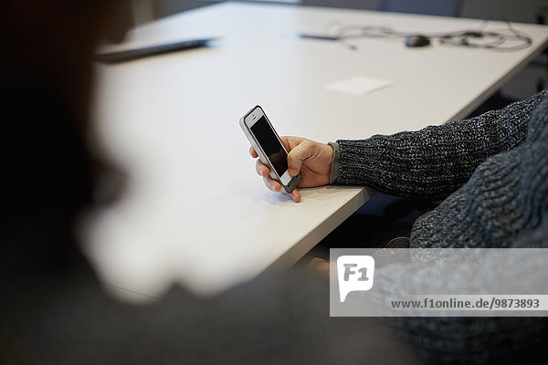 Ein Mann  der an einem Tisch in einem Büro sitzt und sein Smartphone benutzt.