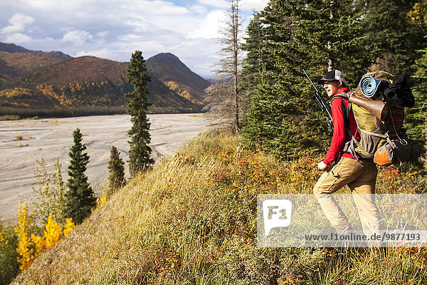 Hunter with backpack and rifle stands on a riverbank overlooking a braided river during autumn  Southcentral Alaska