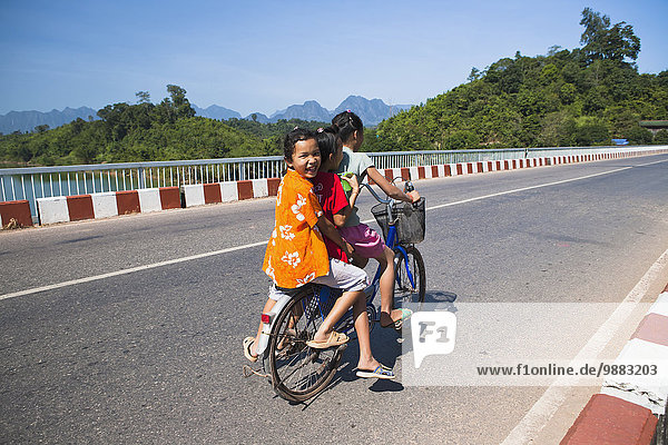 'Young girls ride a bike together over a bridge; The Bak  Laos'