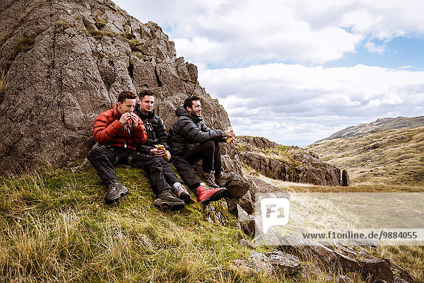 Three young male hiking friends eating sandwiches  The Lake District  Cumbria  UK